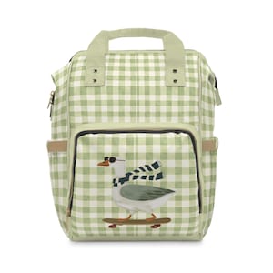 Pode incluir: Uma mochila com padrão xadrez verde sálvia e branco, apresentando um gráfico de um ganso branco com óculos de sol e um cachecol às riscas a andar de skate. A mochila inclui uma pega superior, dois bolsos laterais e um compartimento frontal.