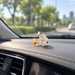 May include: A small white and brown spotted cat figurine sitting on a car dashboard. The cat has a painted face with whiskers and is holding a miniature clear plastic cup filled with an orange liquid and ice, featuring a thin black straw.