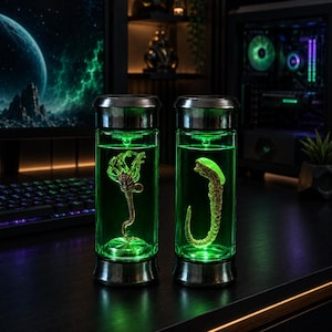May include: Two cylindrical glass containers with metallic silver caps and bases, each filled with a vibrant green liquid. Inside the left container is a tan facehugger figure, and inside the right container is a tan chestburster figure.