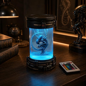 May include: A cylindrical lamp featuring a black base and top with a clear glass center containing a dark blue alien creature figurine. The lamp emits a bright blue light and is accompanied by a small rectangular remote control with colored buttons.