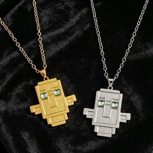 Può includere: Due collane con ciondoli rettangolari in stile pixel, caratterizzati da superfici testurizzate e due piccole pietre quadrate verde chiaro incastonate come occhi. Un ciondolo è color oro e l'altro color argento, entrambi appesi a catene metalliche.