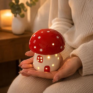 May include: A ceramic mushroom-shaped lamp with a glossy red cap featuring white polka dots and a white base. The base includes cut-out windows and a door, allowing warm light to shine through from the interior, creating a glowing effect.