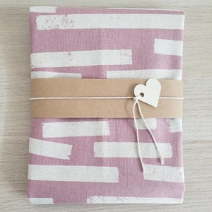 May include: A folded textile with a pink background and white brushstroke design. A brown paper band is wrapped around the textile, secured with a white string and a heart-shaped tag. The textile is likely a fabric item.