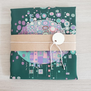 May include: A folded square fabric scarf featuring a dark green background with a central circular pattern of pink, purple, and teal geometric shapes and circles. A tan paper band wraps around the center with a white circular tag attached by string.