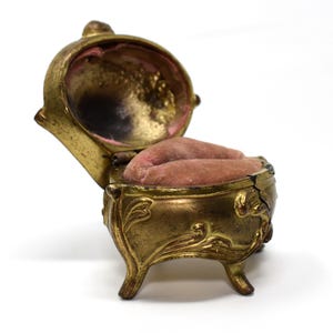 Pode incluir: Uma caixa de joias de metal em tom dourado antigo com tampa articulada aberta. O interior apresenta uma almofada de veludo rosa empoeirado. O exterior tem detalhes ornamentados em relevo e apoia-se em quatro pequenas pernas curvas.