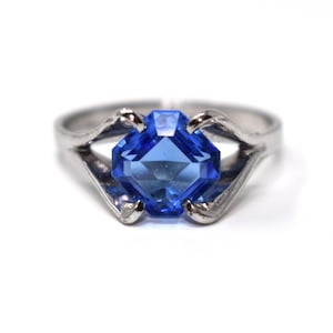 May include: A silver-toned metal ring featuring a central square-cut blue gemstone. The band has a split design that curves upward to hold the stone in a four-prong setting. The metal has a polished finish and the blue stone is faceted.