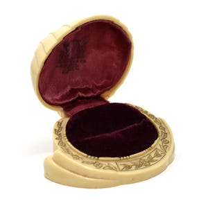 Puede incluir: Un estuche para anillos abierto de color crema con un forro interior de terciopelo burdeos intenso. El exterior presenta un borde con motivos florales tallados y un logotipo oscuro impreso en la tapa interior. La base es curva.