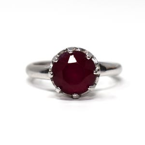 May include: A silver-toned ring featuring a round, deep red faceted gemstone set in a prong setting. The band is smooth and polished, with the circular stone centered prominently in the design, showcasing a dark crimson hue and reflective facets.
