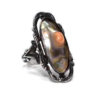 May include: An oval-shaped ring featuring a dark, oxidized silver setting with intricate scrollwork. The center stone is an elongated, iridescent abalone shell inlay with a small, smooth, light orange circular accent piece set within the shell.