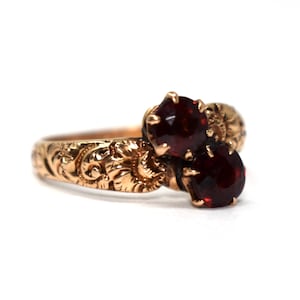 May include: A gold ring featuring two round, deep red garnet gemstones set in a bypass style. The band is crafted from yellow gold and displays an intricate, embossed floral and scrollwork pattern that extends along the entire visible shank.