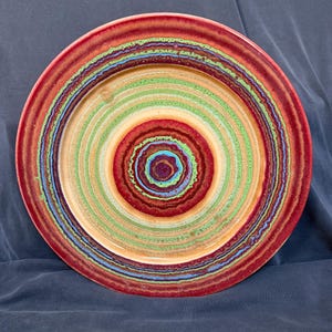 May include: A round ceramic plate featuring a vibrant, concentric ring pattern. The design consists of alternating bands of deep red, bright green, light tan, and blue, creating a textured, multi-colored appearance across the entire surface.