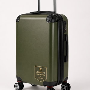 May include: An olive green hard-shell carry-on suitcase with a retractable black handle, four dual-spinner wheels, and a black zipper closure. A rectangular gold-bordered patch on the front displays an airplane icon and the text JOURNEY'S MADE BETTER EST. 2026.