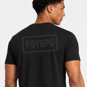 May include: A black short-sleeved t-shirt featuring a rectangular graphic on the back. Inside the rectangle, the word PSYOPS is printed in a dark gray, sans-serif font. The design is centered on the upper back of the garment.