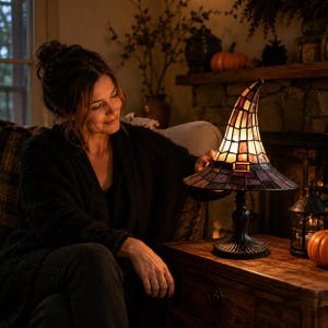 May include: A stained glass table lamp shaped like a pointed witch hat with a black base and a band featuring a buckle detail. The lamp shade is constructed from multiple glass panels in shades of amber, brown, and purple, emitting a warm glow.