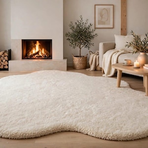 May include: A large, off-white area rug with an irregular, organic, cloud-like shape. The rug features a thick, plush, high-pile texture that covers the floor surface, providing a soft and dense appearance throughout its entire rounded perimeter.