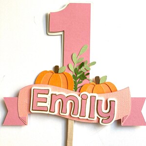 May include: A pink and white number one cake topper with two orange pumpkins, green leaves, and the name "Emily" on a banner. The topper is on a wooden stick, perfect for a first birthday celebration.