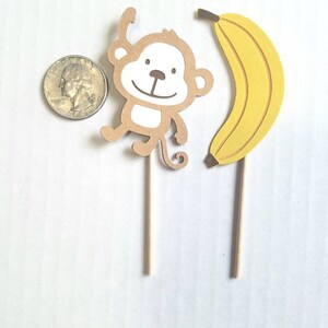 May include: Two brown and yellow paper cupcake toppers, one with a monkey design and the other with a banana design. The monkey is brown with a white face and is holding its arms up. The banana is yellow and curved. Both toppers are attached to wooden sticks.