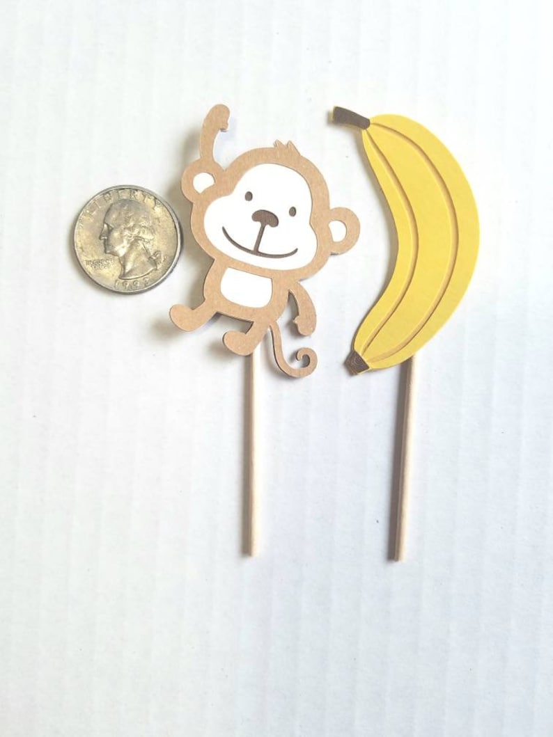 May include: Two brown and yellow paper cupcake toppers, one with a monkey design and the other with a banana design. The monkey is brown with a white face and is holding its arms up. The banana is yellow and curved. Both toppers are attached to wooden sticks.