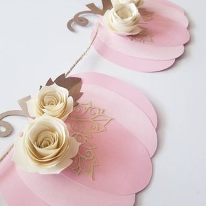 May include: A pink and gold pumpkin garland with white paper flowers. The garland is made of paper and is strung together with twine.