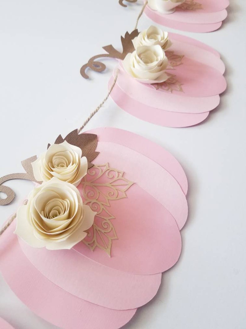 May include: A pink and gold pumpkin garland with white paper flowers. The garland is made of paper and is strung together with twine.