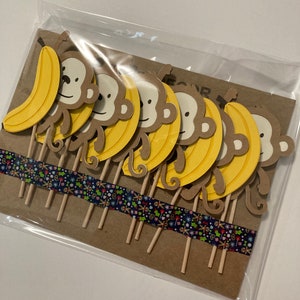 May include: A set of 8 brown monkey cupcake toppers with yellow bananas. The toppers are on wooden sticks and are packaged in a clear plastic bag.