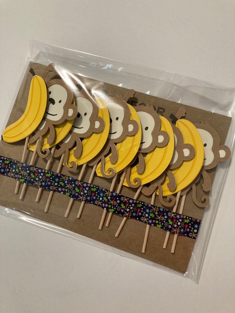 May include: A set of 8 brown monkey cupcake toppers with yellow bananas. The toppers are on wooden sticks and are packaged in a clear plastic bag.
