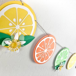 May include: A paper garland with alternating yellow lemon, orange, and green lime slices. Each slice has a white center and is decorated with a white flower with a yellow center and green leaves.