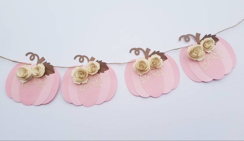 May include: A pink and brown paper pumpkin garland with white paper flowers. The garland is strung together with twine.