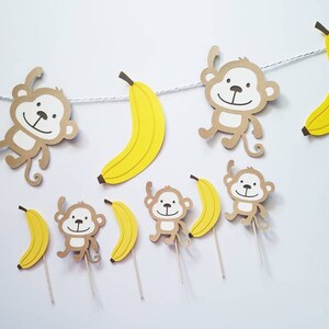 May include: A brown paper banner with yellow paper bananas and monkeys hanging from it. The monkeys have brown fur and white faces. There are also individual cupcake toppers with monkeys and bananas.