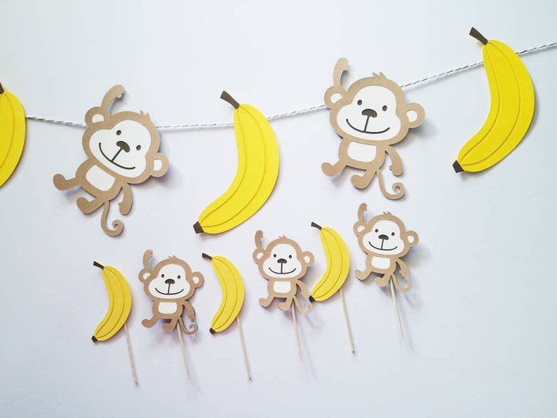 May include: A brown paper banner with yellow paper bananas and monkeys hanging from it. The monkeys have brown fur and white faces. There are also individual cupcake toppers with monkeys and bananas.