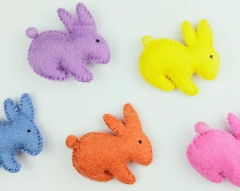 May include: Five felt bunny rabbits in different colors: purple, yellow, blue, orange, and pink. The bunnies are all facing the right side of the image.