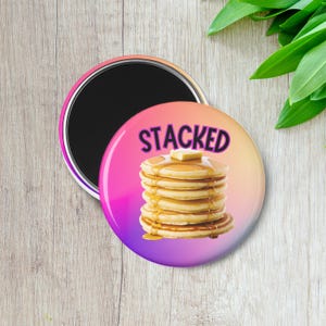 May include: A round magnet with a stack of pancakes, butter, and syrup. The word "STACKED" is above the pancakes. The magnet has a gradient background of pink, orange, and purple.