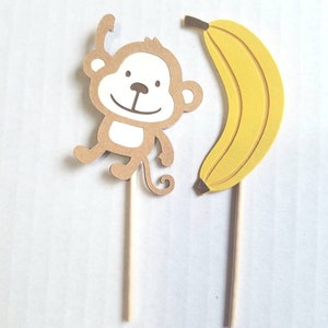 May include: Two brown and yellow paper cupcake toppers, one with a monkey design and the other with a banana design. The toppers are attached to wooden sticks.