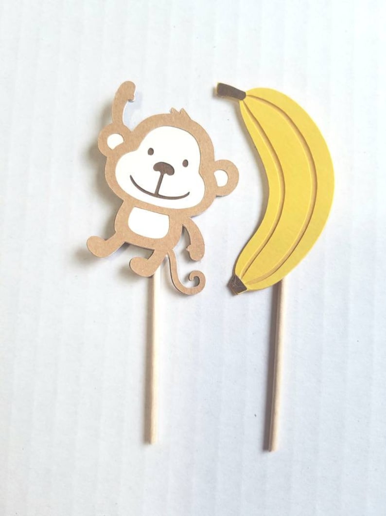 May include: Two brown and yellow paper cupcake toppers, one with a monkey design and the other with a banana design. The toppers are attached to wooden sticks.