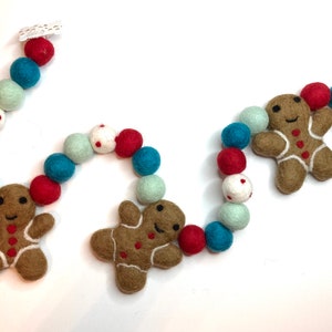May include: A festive felt garland featuring gingerbread men and colorful felt balls in red, teal, white, and mint green. The garland is designed for holiday or Christmas decorations, with a cheerful and handmade aesthetic.
