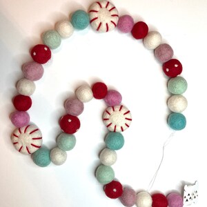 May include: A festive garland featuring a mix of felt balls in various colors, including red, pink, white, and teal. The garland also includes peppermint-shaped ornaments with red stripes. A small wooden accent with a bear design is attached to the end.