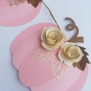 May include: A pink paper pumpkin garland with brown paper leaves and cream paper flowers. The garland is strung together with twine.