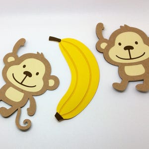 May include: Two brown paper monkey cutouts with smiling faces and a yellow paper banana cutout.