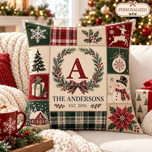 May include: A square throw pillow featuring a patchwork design with Christmas motifs including a central wreath with the letter A, the text THE ANDERSONS EST. 2026, a snowman, a reindeer, a Christmas tree, a gift box, a house, and poinsettia.