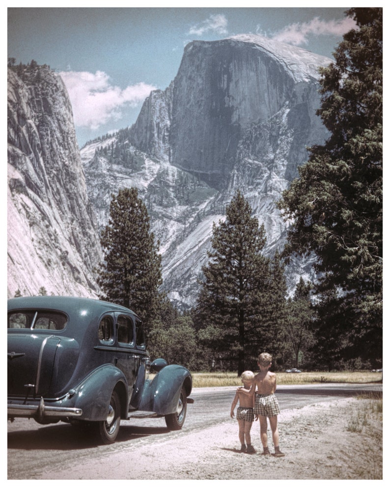 May include: Vintage photograph of a teal-colored classic car parked on a dirt road with two young children standing nearby. The background features a large mountain and tall trees under a blue sky.