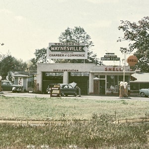 May include: Vintage photograph of a Shell gas station in Waynesville, with classic cars and signage. The sign reads "Welcome to Waynesville" and "Shell Lubrication". A sign indicates directions to Cherokee and Murphy.