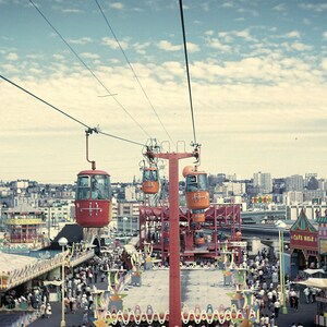 May include: An aerial view of an amusement park with a red and orange gondola lift system. The sky is blue with scattered clouds. Below, crowds of people walk around the park, with various rides and buildings visible.