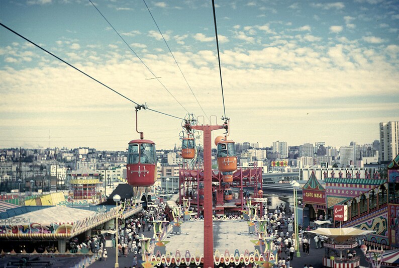 May include: An aerial view of an amusement park with a red and orange gondola lift system. The sky is blue with scattered clouds. Below, crowds of people walk around the park, with various rides and buildings visible.