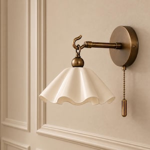 Peut inclure: Une applique murale composée d'un bras en métal couleur laiton et d'une platine circulaire. Un abat-jour blanc en forme de cloche à bords ondulés est suspendu à un crochet. Une chaînette en laiton avec un embout cylindrique est fixée.