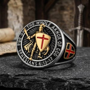 Könnte beinhalten: Ein silberfarbener Ring mit einem goldfarbenen Ritter, der einen Schild mit einem roten Kreuz hält. Der kreisförmige Rand ist mit dem Text PUT ON THE WHOLE ARMOR OF GOD EPHESIANS 6:13-17 graviert. Ein kleiner Schild mit rotem Kreuz ist seitlich.