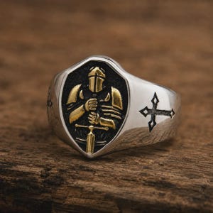Könnte beinhalten: Ein silberfarbener Ring mit einem schildförmigen Mittelstück, das eine goldfarbene Ritterfigur mit Schwert vor einem dunklen, strukturierten Hintergrund zeigt. Die Seiten des Rings sind mit einem schwarzen Kreuzemblem auf jeder Seite verziert.