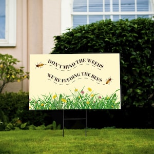 May include: A yellow sign with brown text that reads "Don't mind the weeds, we're feeding the bees". The sign has a green grass border with yellow flowers and bees flying around it.