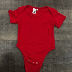 May include: A red baby onesie with short sleeves and snap closures. The onesie has a white tag with the letters "JB" on it.