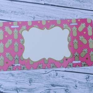 May include: Pink and gold license plate with a pineapple pattern and a blank white space in the center. The license plate has two holes on the top and two holes on the bottom.