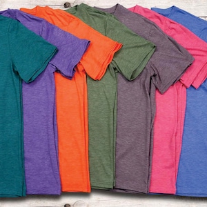 May include: A collection of seven short-sleeved t-shirts in various colors, including teal, purple, orange, green, grey, pink, and blue. The shirts are arranged in a row, showcasing their different hues and textures. The background is a light-colored wooden surface.